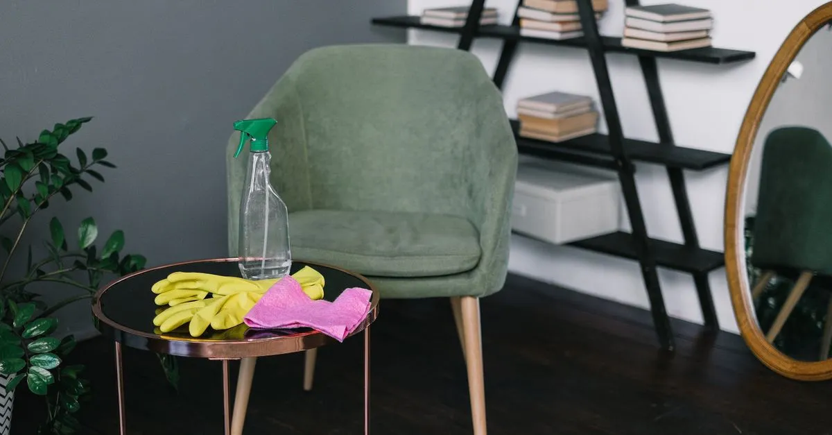 Yellow latex gloves placed near spray detergent bottle and rag on round table against comfortable chair in living room i